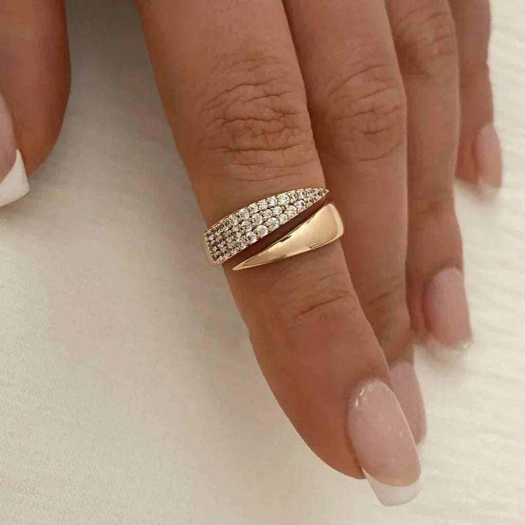 A hand with manicured nails wears a stylish gold ring featuring a twisted design that wraps around the finger. One side boasts a polished finish, while the other is encrusted with sparkling pavé diamonds.