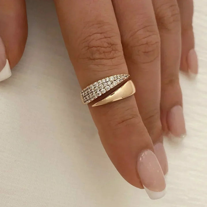 A hand with manicured nails wears a stylish gold ring featuring a twisted design that wraps around the finger. One side boasts a polished finish, while the other is encrusted with sparkling pavé diamonds.