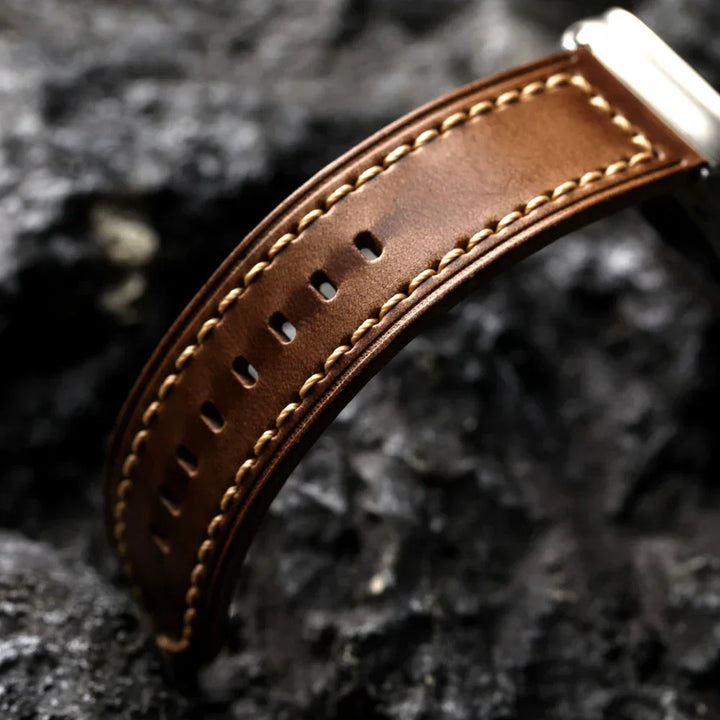 Close-up of a handcrafted brown cowhide leather watch strap with beige stitching and multiple holes, set against a textured, dark gray rocky background.