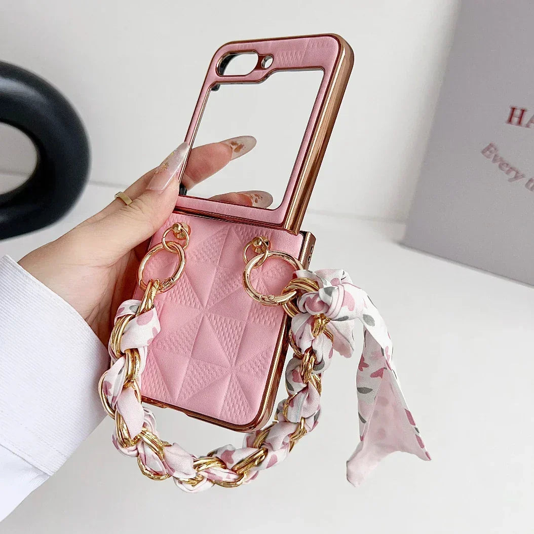 A hand holds a pink Samsung Galaxy Z Flip premium leather case with a quilted pattern, built-in mirror, shockproof protection, and a decorative chain handle wrapped with a floral scarf. A white wall and partially visible book are in the background.