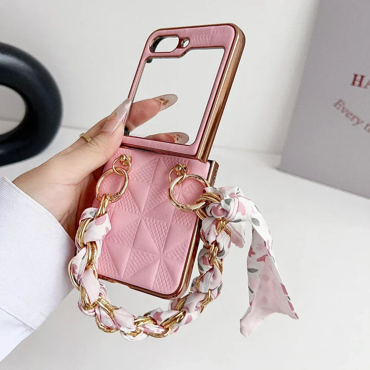 A hand holds a pink Samsung Galaxy Z Flip premium leather case with a quilted pattern, built-in mirror, shockproof protection, and a decorative chain handle wrapped with a floral scarf. A white wall and partially visible book are in the background.