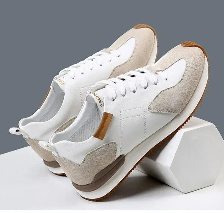 A pair of stylish white sneakers with top-grain leather, light beige suede accents, and brown rubber soles offer excellent arch support, displayed on white geometric blocks against a gray background.