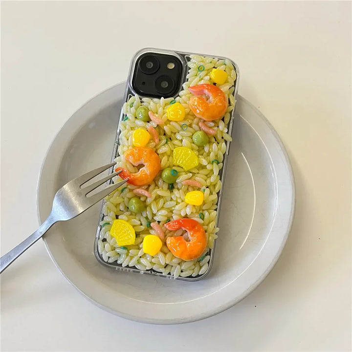 A smartphone case with 3D detailing mimics a plate of rice with shrimp, corn, and peas on a white plate. A fork rests on the case, enhancing the food-like illusion while offering rugged phone protection.