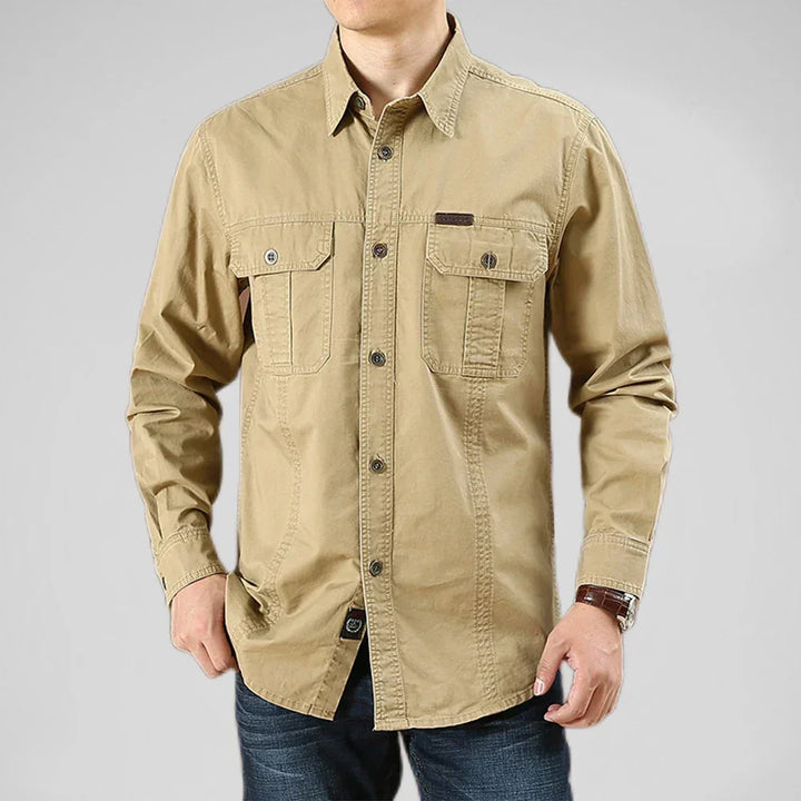 A man wearing a light brown 100% cotton shirt with two chest pockets and dark jeans, standing against a plain light gray background. His face is not visible.