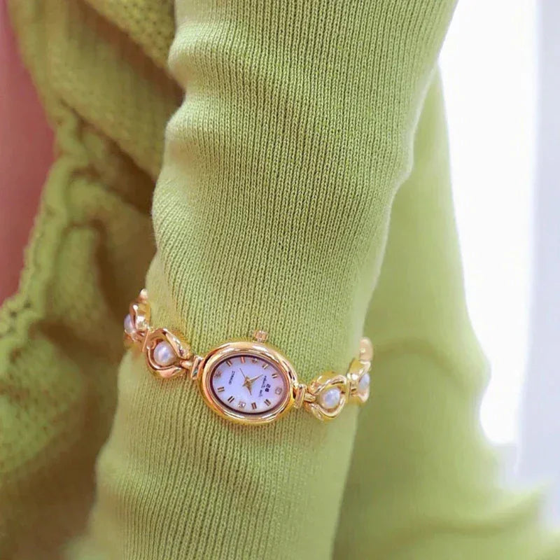 A gold bracelet watch with precision quartz movement and pearl accents is worn over a light green, ruffled long-sleeve top. The small, oval watch face displays Roman numerals.