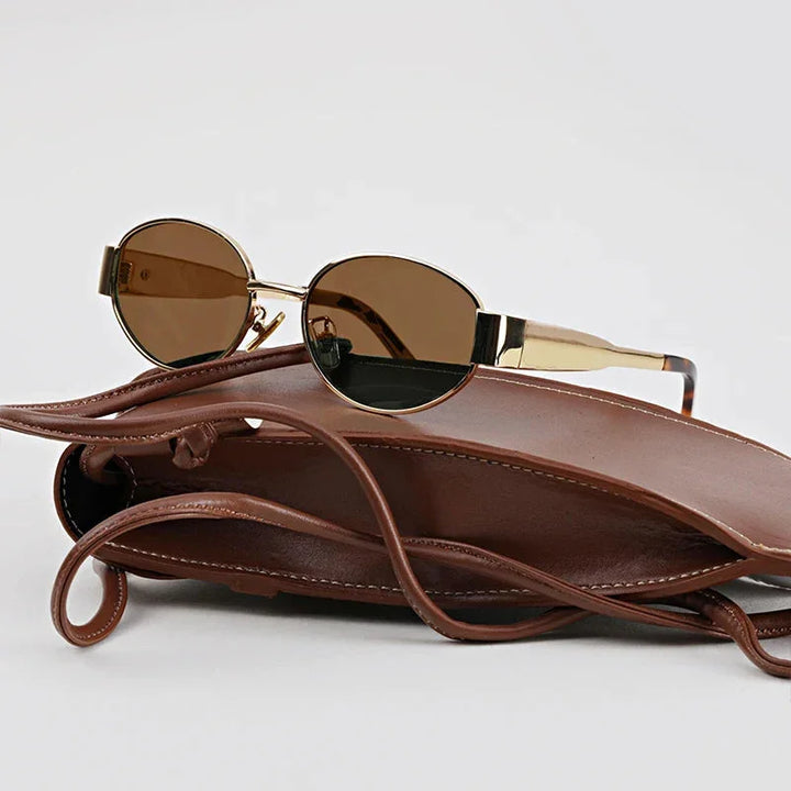 Round, gold-framed, vintage-inspired sunglasses with dark lenses and UV400 protection rest on top of an open brown leather handbag with long straps, set against a plain light gray background.