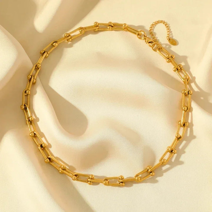 A gold statement chain necklace with elongated links and small round beads, crafted from durable 316L stainless steel and displayed on a soft cream-colored fabric background.