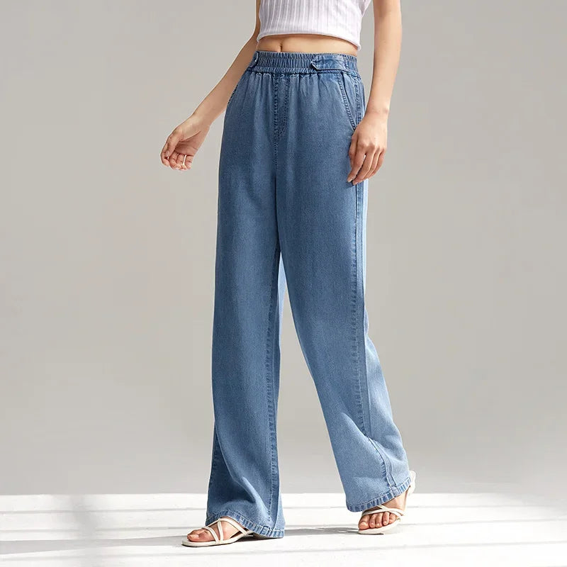 A person wearing light blue, wide-leg, breathable denim pants with an elastic waistband, a white sleeveless crop top, and white sandals stands against a plain, light background. Only the lower half of their body is visible.