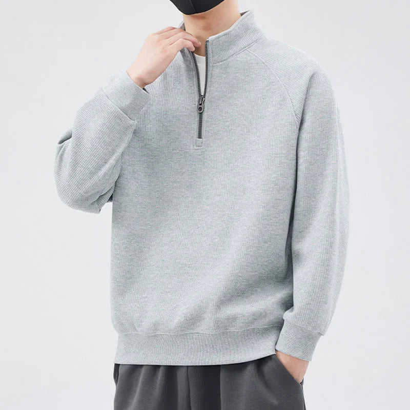 A person wearing a light gray pullover sweater with a half-zip closure and dark pants stands against a plain light background. The relaxed fit sweater features a stand-up collar, and the person has one hand raised to their face with a black mask.