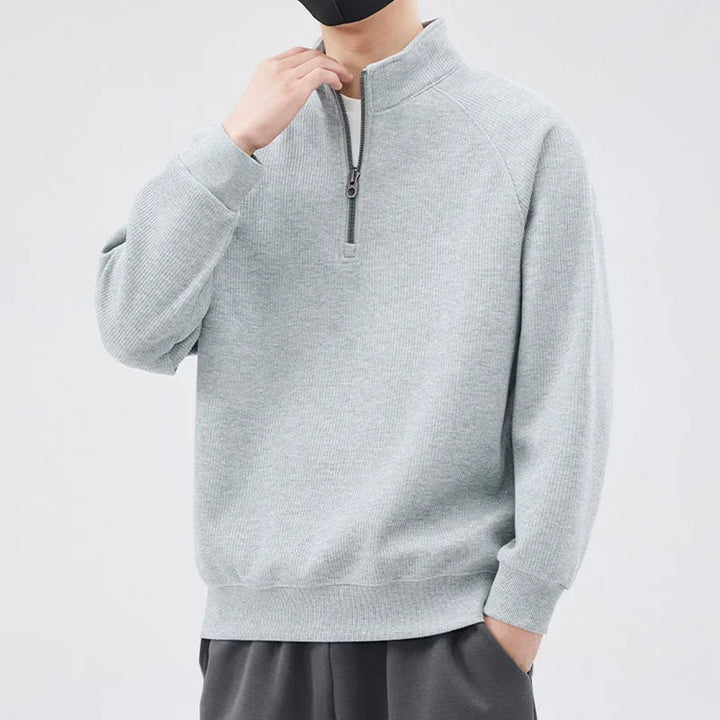 A person wearing a light gray pullover sweater with a half-zip closure and dark pants stands against a plain light background. The relaxed fit sweater features a stand-up collar, and the person has one hand raised to their face with a black mask.