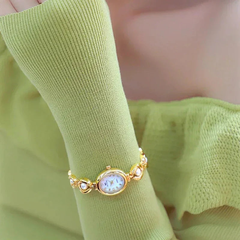 A person wearing a light green long-sleeved top displays a gold wristwatch with pearl accents. The watch features an oval face with a white dial, precision quartz movement, and is stylishly worn over the sleeve.