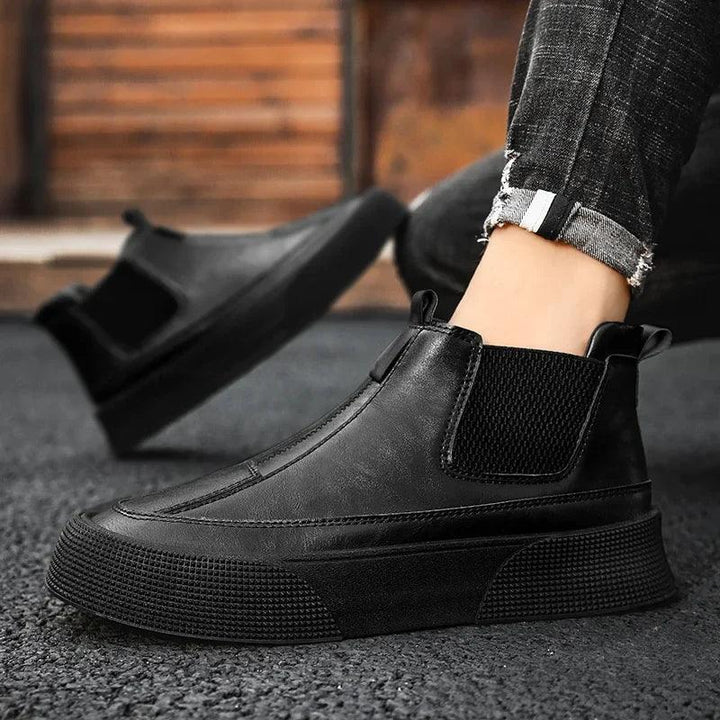 A person wearing black vintage high-tops with thick soles, elastic side panels, and a cushioned footbed stands on a textured outdoor surface. Another foot in the same shoe appears in the background. The person is wearing rolled-up jeans.