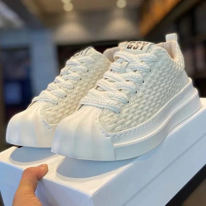 A hand holds a box displaying white woven upper sneakers with thick, cushioned soles and “GSY” printed on the tongue, set against a blurred indoor background.