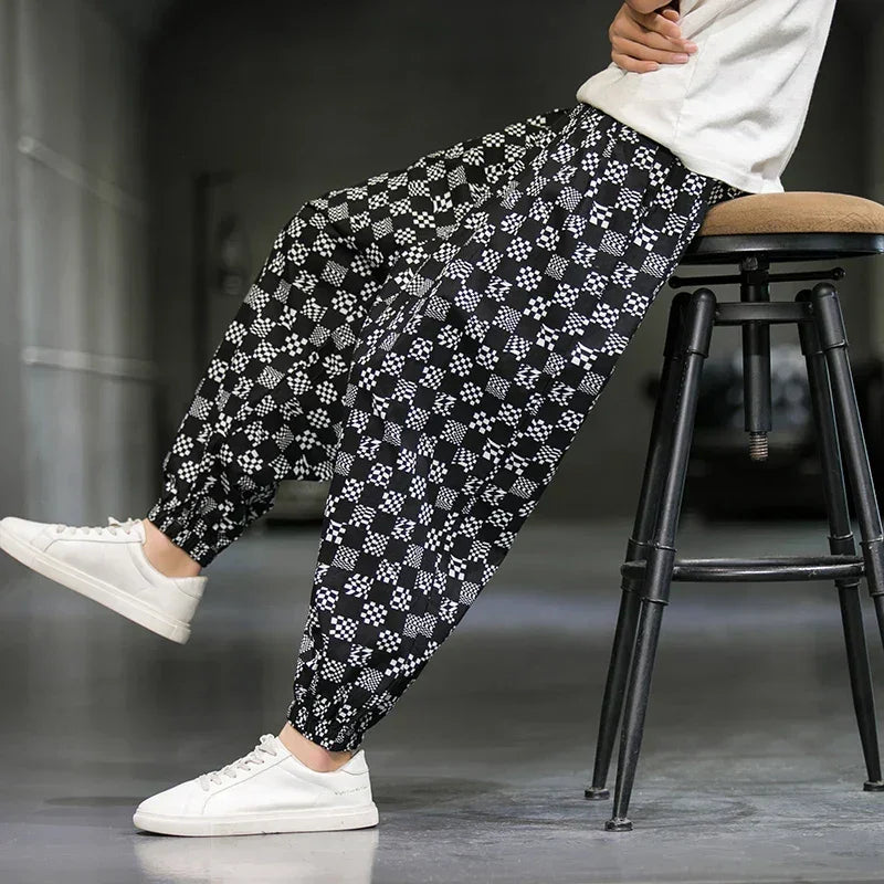 A person sits sideways on a stool wearing black cotton joggers with a white geometric pattern, a white shirt, and white sneakers—making a subtle fashion statement against a minimal, blurred background.