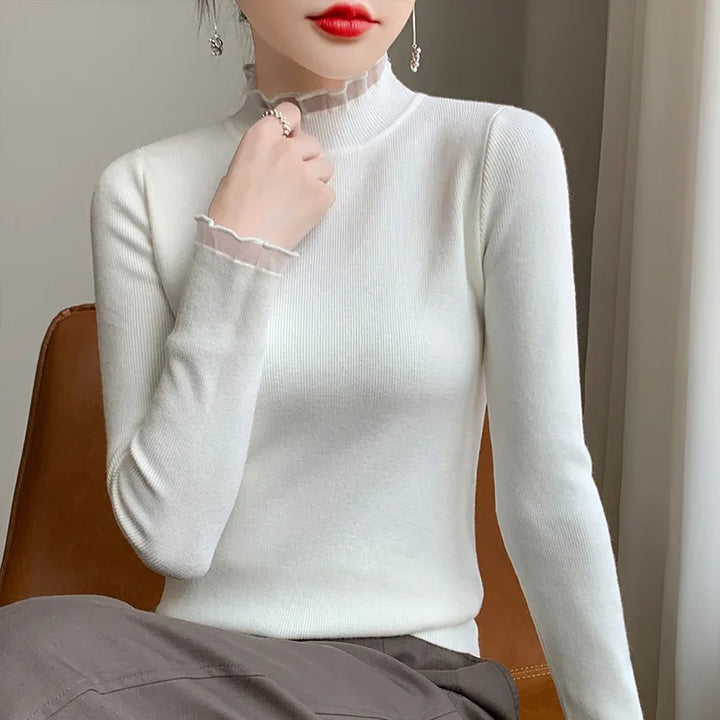A woman wears a fitted cashmere blend white turtleneck sweater with sheer ruffled edges on the neck and cuffs, sitting on a brown chair, her hand touching her neck. She has red lipstick and sits near a window.