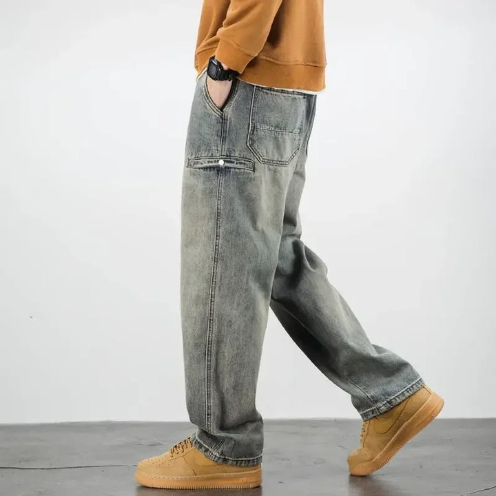 Person wearing a brown sweatshirt, classic fit stone washed jeans, and tan sneakers, standing sideways with hands in pockets against a plain light gray background.