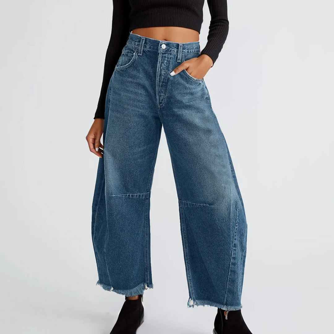 Woman wearing high-waisted, relaxed fit blue jeans with frayed hems, a black long-sleeve crop top, and black ankle boots. One hand is in her pocket against a plain light background—perfect for styling your favorite summer jean look.
