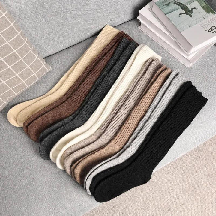 A row of ribbed, breathable socks in various neutral colors—cream, beige, brown, gray, and black—made from a soft Cashmere cotton blend, neatly arranged on a light gray couch next to a checkered pillow and books.
