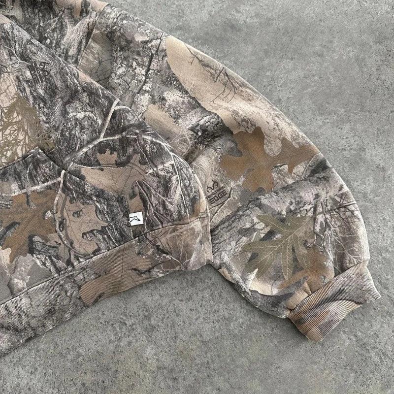 A close-up of a camo pattern sweatshirt sleeve in earthy tones, crafted from a soft cotton blend and resting on a gray concrete surface, with realistic leaf and tree branch designs.