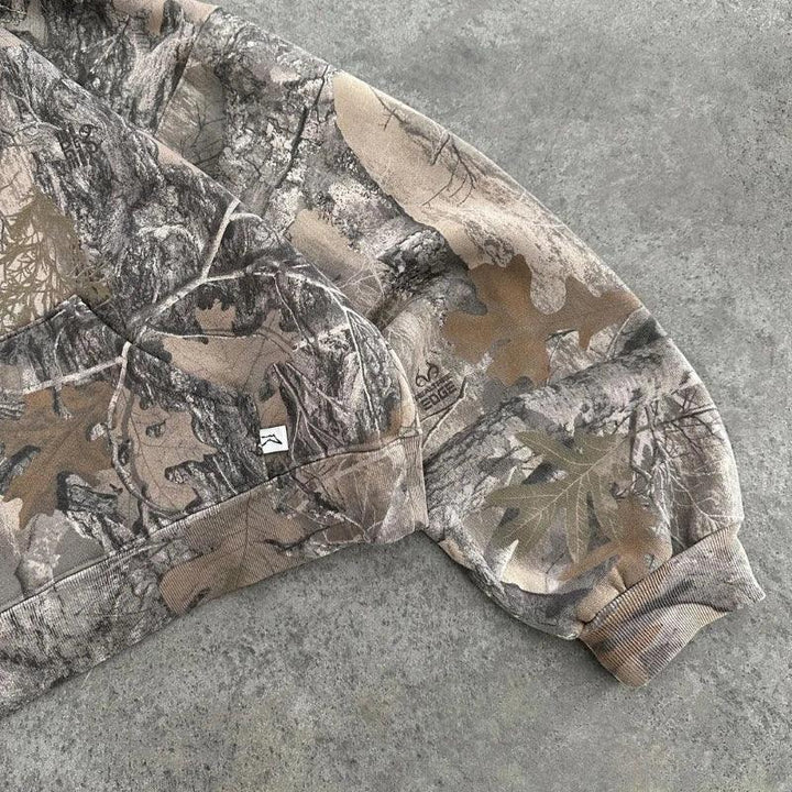 A close-up of a camo pattern sweatshirt sleeve in earthy tones, crafted from a soft cotton blend and resting on a gray concrete surface, with realistic leaf and tree branch designs.