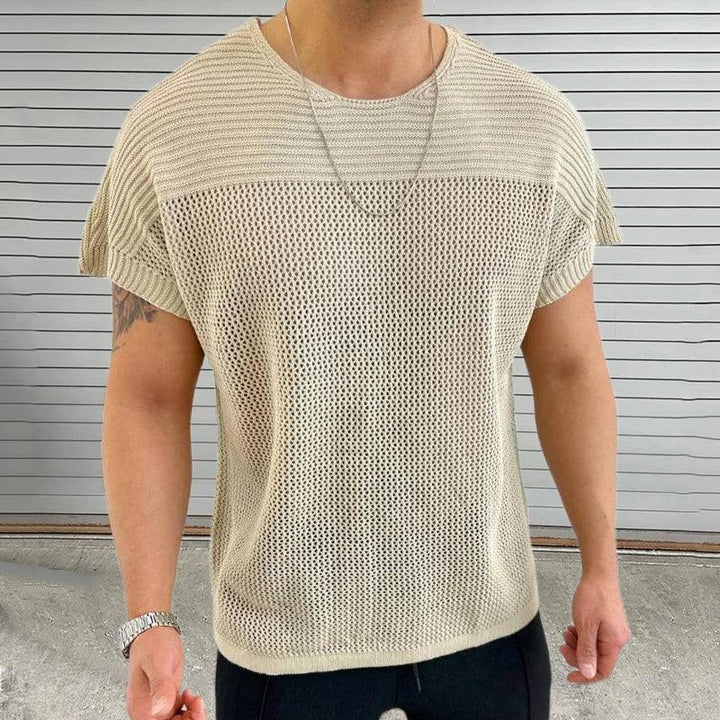 A man wearing a light beige, short-sleeve mesh knit tee with a breathable design and a silver necklace stands in front of a gray, horizontal metal door. Only his upper body is visible.