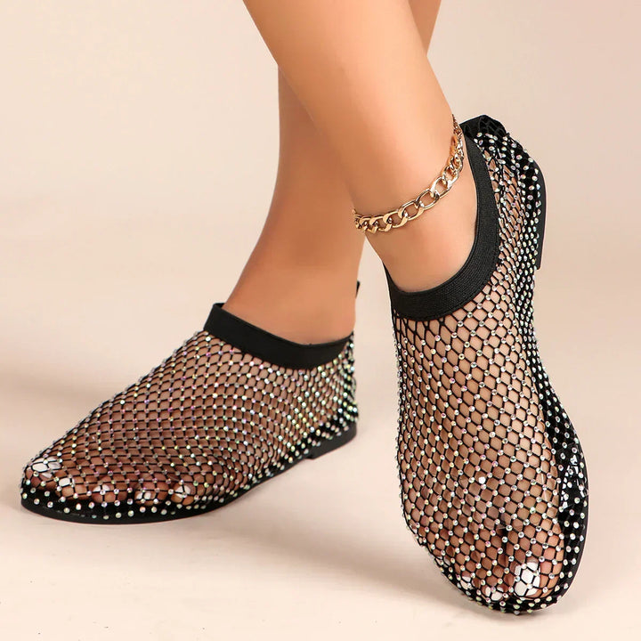 A person wearing black mesh overlay shoes with rhinestone accents and a gold chain anklet, standing on a neutral background.