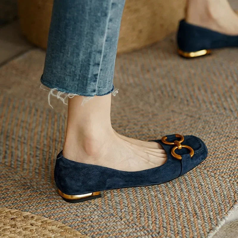 A person wearing frayed blue jeans and navy blue suede flats with cushioned insoles and gold metallic accents stands on a woven rug.