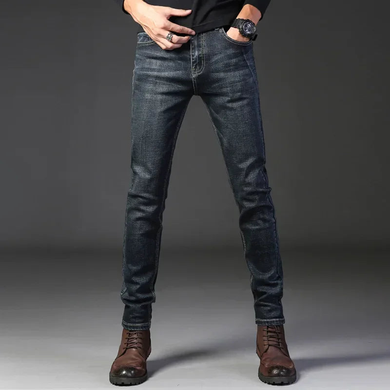 A person wearing dark blue slim-fit jeans crafted from stretch denim, a black long-sleeve shirt, brown lace-up boots, and a wristwatch stands against a plain dark background. Their head is not visible in the image.