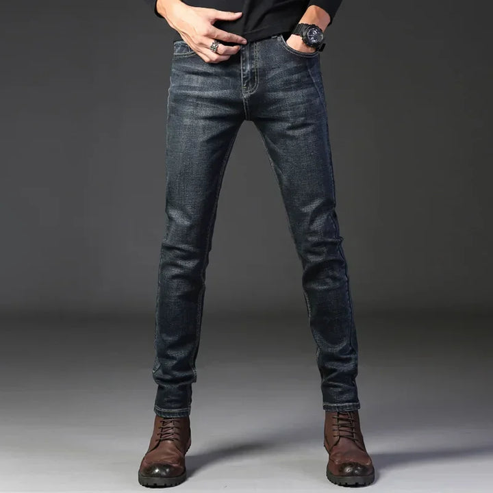 A person wearing dark blue slim-fit jeans crafted from stretch denim, a black long-sleeve shirt, brown lace-up boots, and a wristwatch stands against a plain dark background. Their head is not visible in the image.
