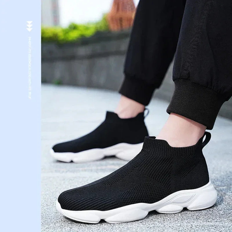 A person wearing lightweight black slip-on sneakers with a non-slip sole and thick white base stands on a paved surface outdoors. Only their lower legs and feet are visible, dressed in black pants.