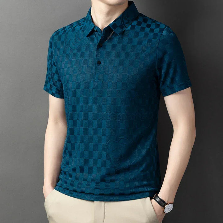 A man wearing a short-sleeved, dark teal checkerboard pattern polo shirt in a tailored fit with beige pants, a black watch, and his hands in his pockets, standing against a plain gray background.