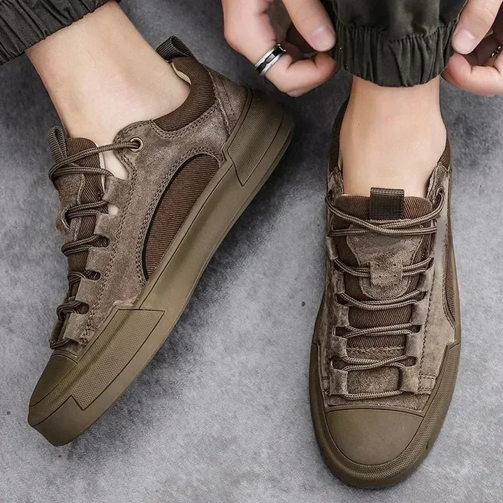 A person wearing brown lace-up sneakers with textured suede and mesh panels, featuring a cushioned insole, stands on a gray surface while adjusting the hem of olive green pants.