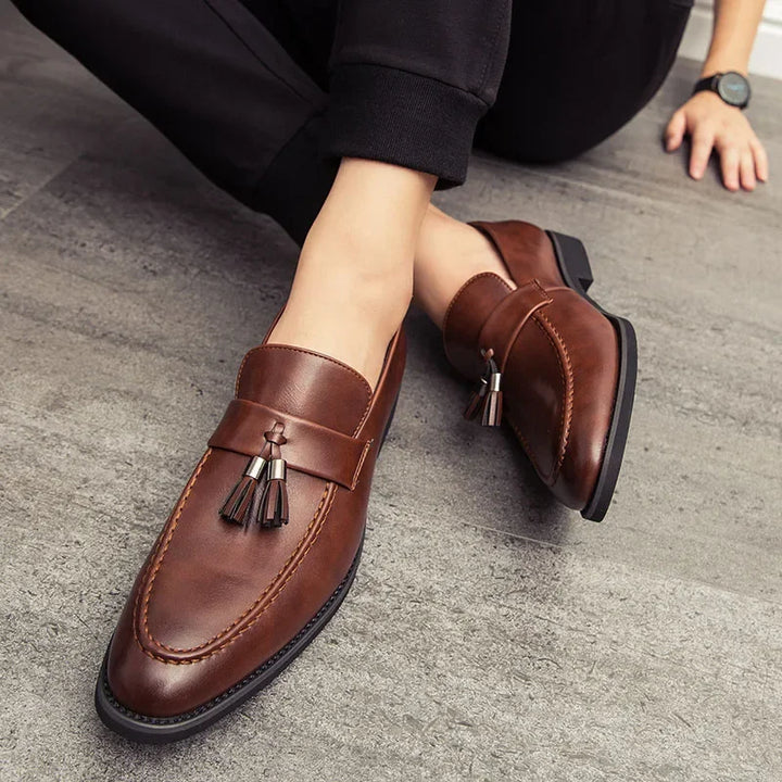 Person wearing polished leather tassel loafers sits on a gray tiled floor. Only the lower legs and one hand are visible, with the person dressed in black pants for a minimalistic shoes look.