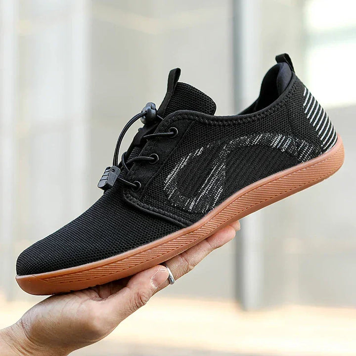 A hand holding a black athletic shoe with a cushioned sole, brown bottom, white and gray stripe accents, mesh upper, and elastic lace closure, shown against a blurred urban background.