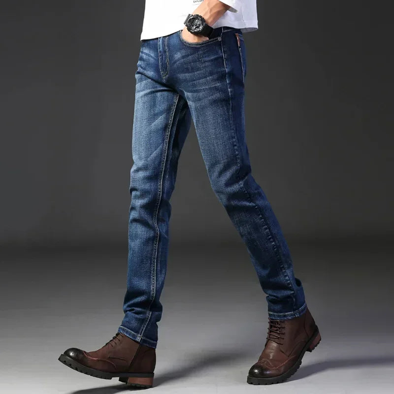 A person wearing blue stretch denim slim-fit jeans, a white shirt, brown lace-up boots, and a black wristwatch stands against a dark gray background. Only the lower half of the body is visible.