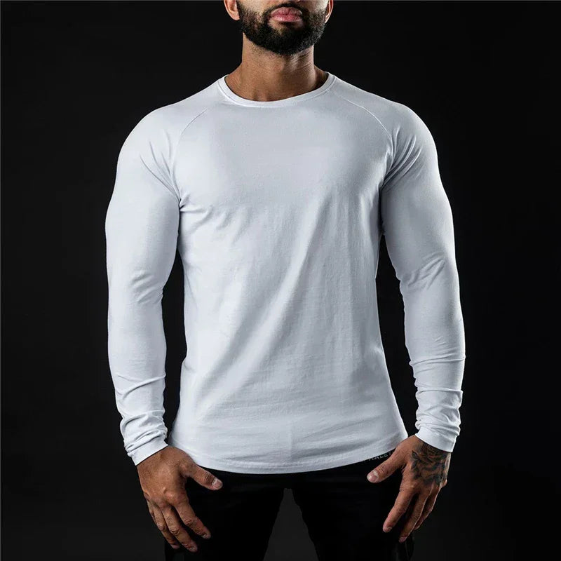 A man wearing a long sleeve fitted shirt stands against a black background, his hands resting by his sides. His head is slightly tilted down and his face is partially out of frame.