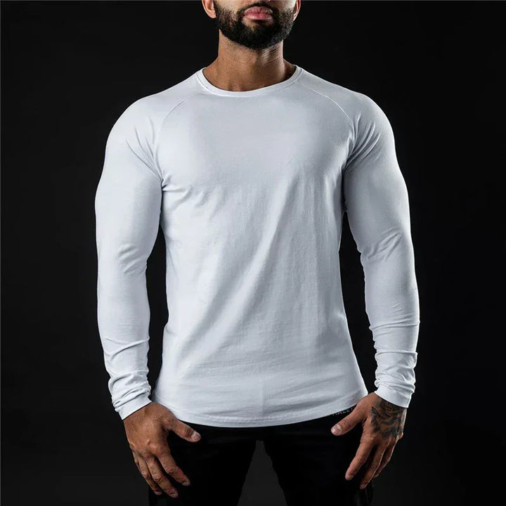 A man wearing a long sleeve fitted shirt stands against a black background, his hands resting by his sides. His head is slightly tilted down and his face is partially out of frame.
