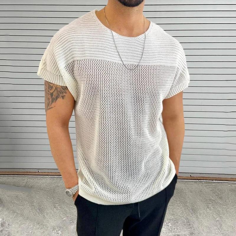 A man wears a white mesh knit tee with a breathable design, black pants, a wristwatch, a silver chain necklace, and shows off an arm tattoo as he stands before a gray horizontal slatted background.