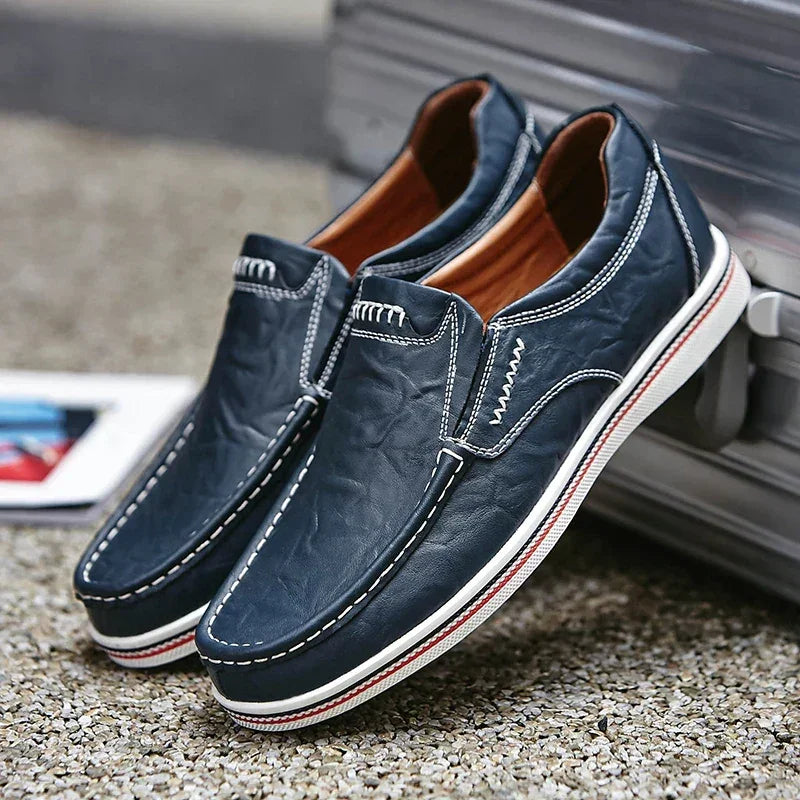 A pair of navy blue boat-style loafers with white stitching and rubber soles, displayed outdoors leaning against a silver suitcase.