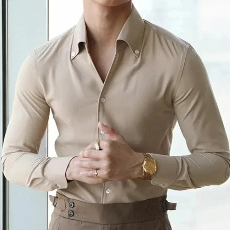 A person wears a slim fit cotton bamboo shirt in beige with dark beige trousers, standing indoors with hands clasped at the waist. A gold watch and a ring complete the look. Their head is not visible in the image.
