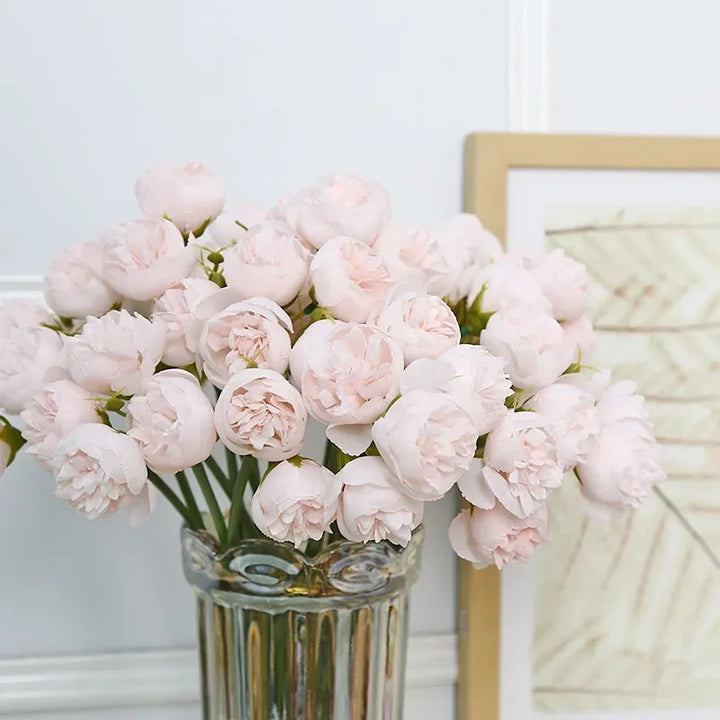 A glass vase filled with light pink peony flowers, featuring realistic faux petals, is placed in front of a framed artwork with neutral tones, against a white paneled wall.