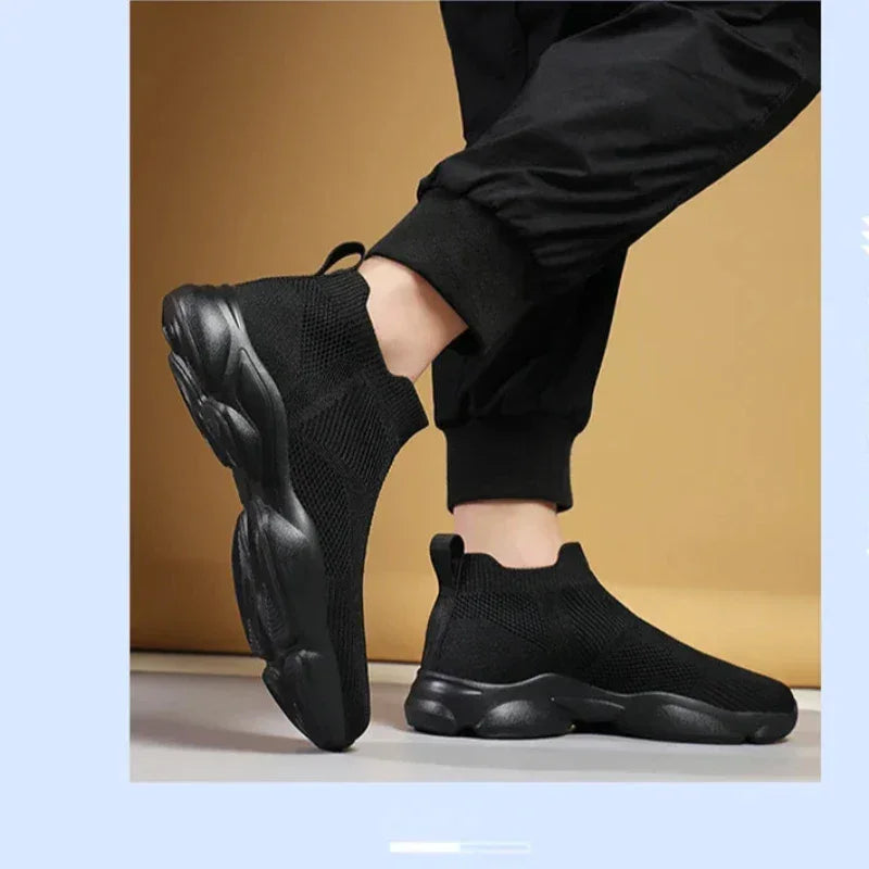 A person wearing black slip-on athletic sneakers with a non-slip sole and black jogger pants stands on a light-colored floor against a beige background. The shoes feature a chunky sole and a breathable knit fabric upper.