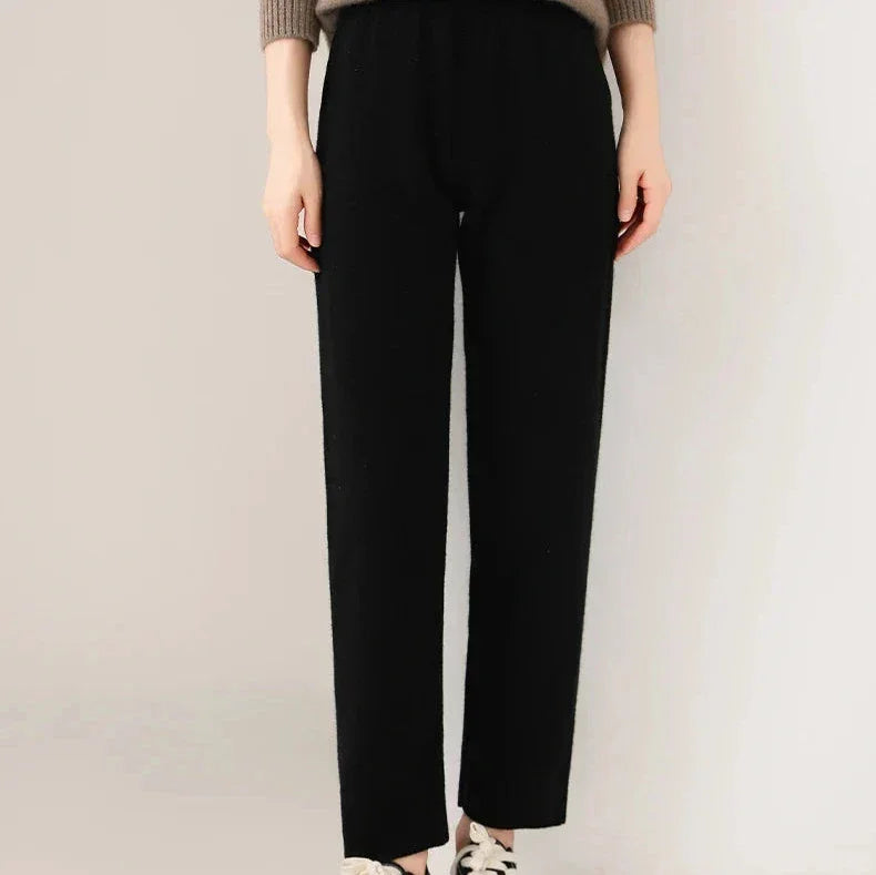 A person wearing black, slim tailored leg pants, a taupe sweater, and white sneakers is standing against a light background. Only the lower half of the body is visible.