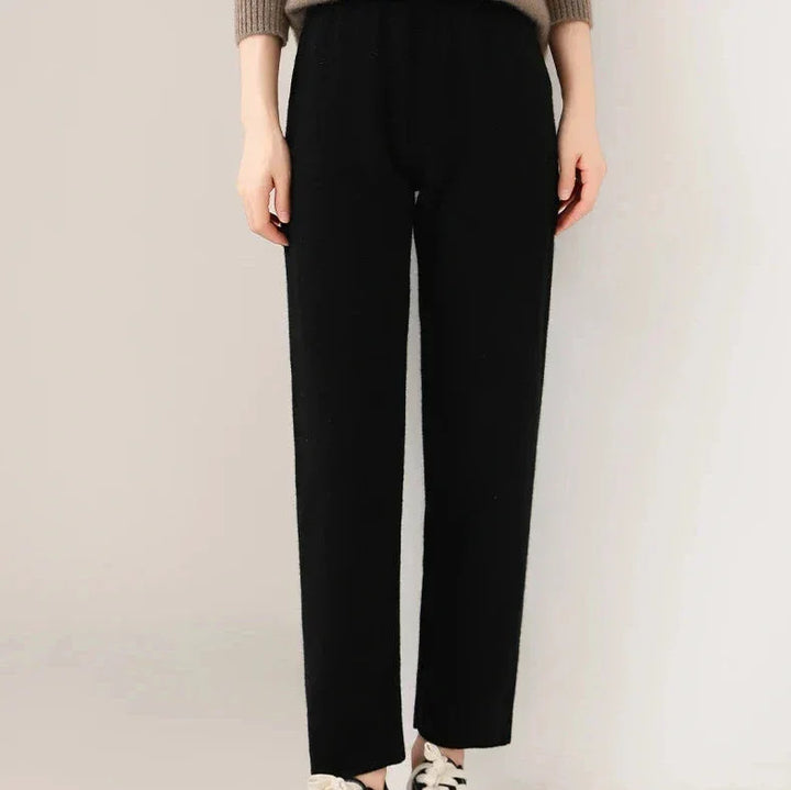 A person wearing black, slim tailored leg pants, a taupe sweater, and white sneakers is standing against a light background. Only the lower half of the body is visible.