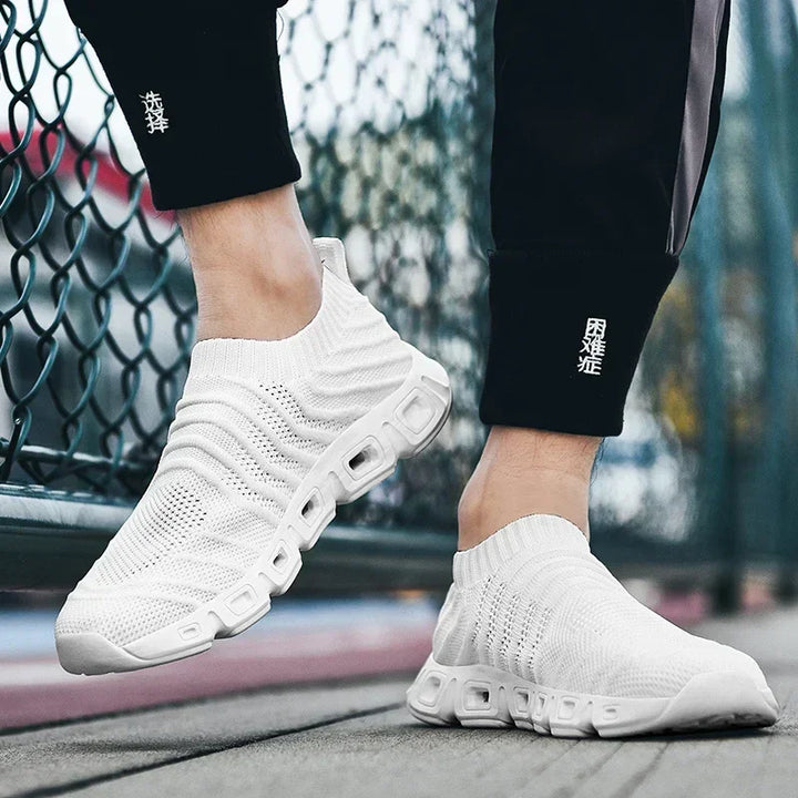 A person wearing white, slip-on athletic sneakers with a sock-like fit and thick, ventilated soles stands near a chain-link fence. The textured knit design complements pants featuring Chinese characters on the lower legs.