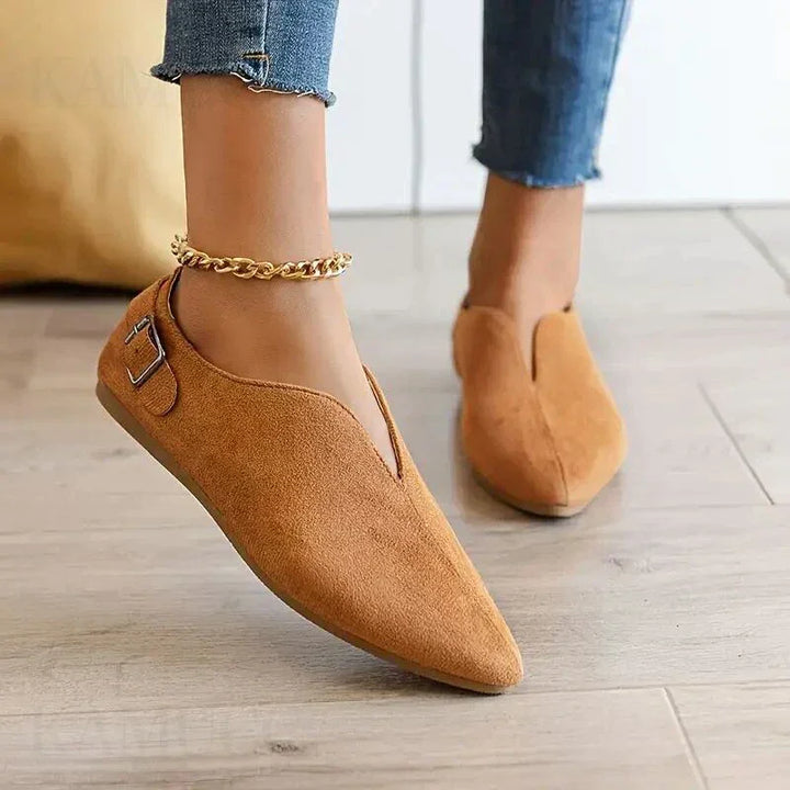 A person wearing tan suede pointed toe shoes with a buckle detail and cushioned insole, paired with rolled-up blue jeans and a gold anklet, stands on a light wooden floor.