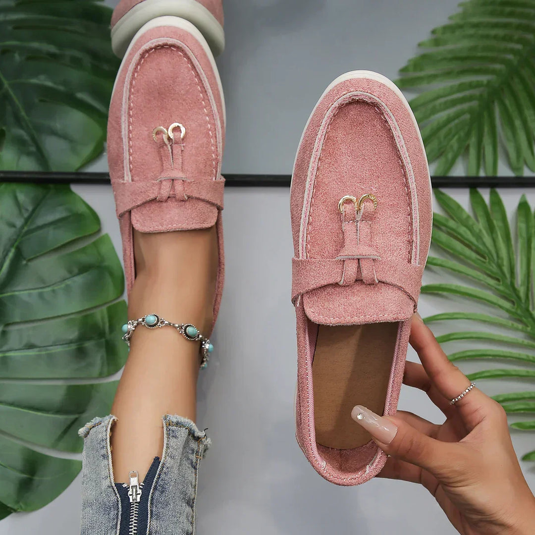 A person wearing a turquoise beaded anklet and ripped jeans tries on chic moccasins in pink suede with white soles, surrounded by green tropical leaves. One loafer is on the foot; the other, held in hand.