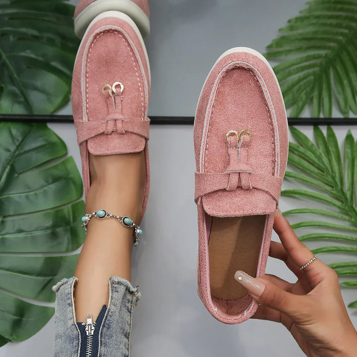 A person wearing a turquoise beaded anklet and ripped jeans tries on chic moccasins in pink suede with white soles, surrounded by green tropical leaves. One loafer is on the foot; the other, held in hand.