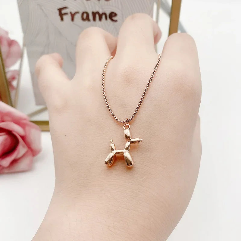 A hand wearing a playful balloon dog necklace with a gold pendant and adjustable chain. Pink roses and a photo frame are softly blurred in the background.