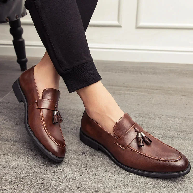 A person wearing polished leather tassel loafers and black pants sits on a chair, showing off the minimalistic shoes on a gray floor with a white wall background.